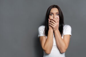 Scared woman covering mouth with hands while posing to camera on gray studio background. Shocked girl close lips with palms, speak no evil concept